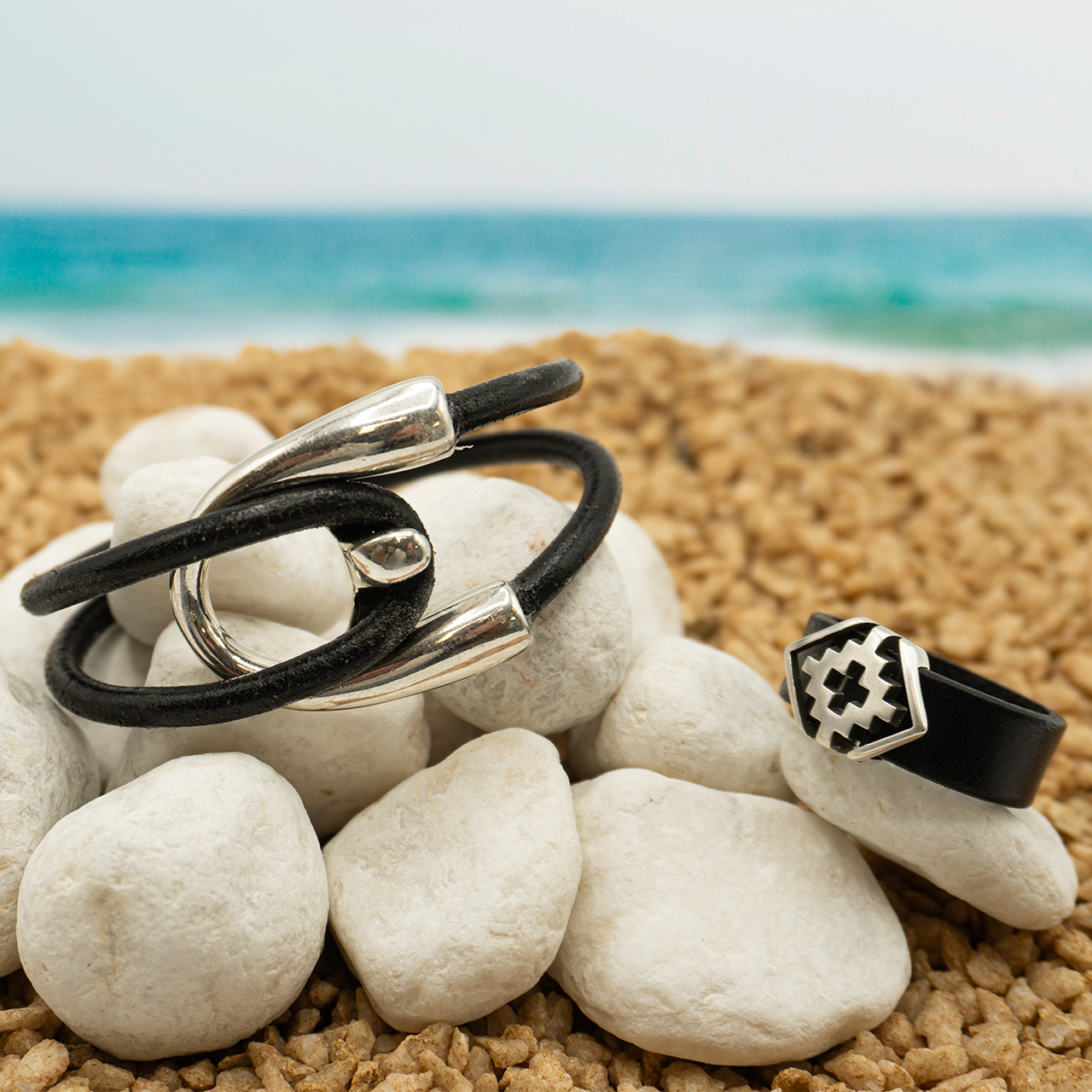 Pulsera de cuero doble y cierre de metal y anillo geométrico sobre piedras blancas en la playa
